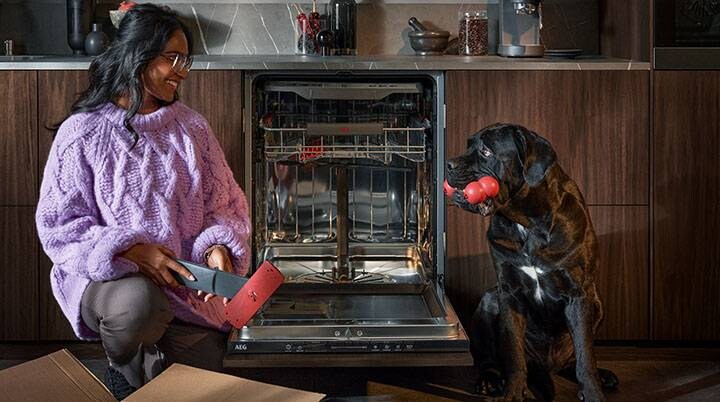 Femme déballant des pièces détachées devant un lave-vaisselle ouvert, tout en souriant à un chien noir.